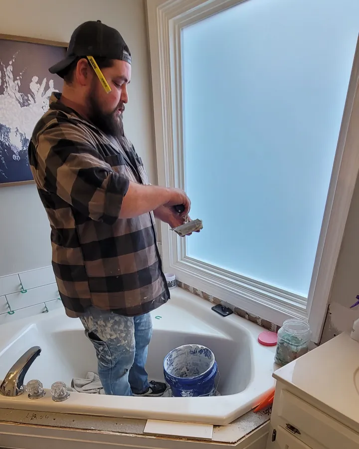 Brandon prepares tile material while standing inside a bathtub during a bathroom remodeling project.