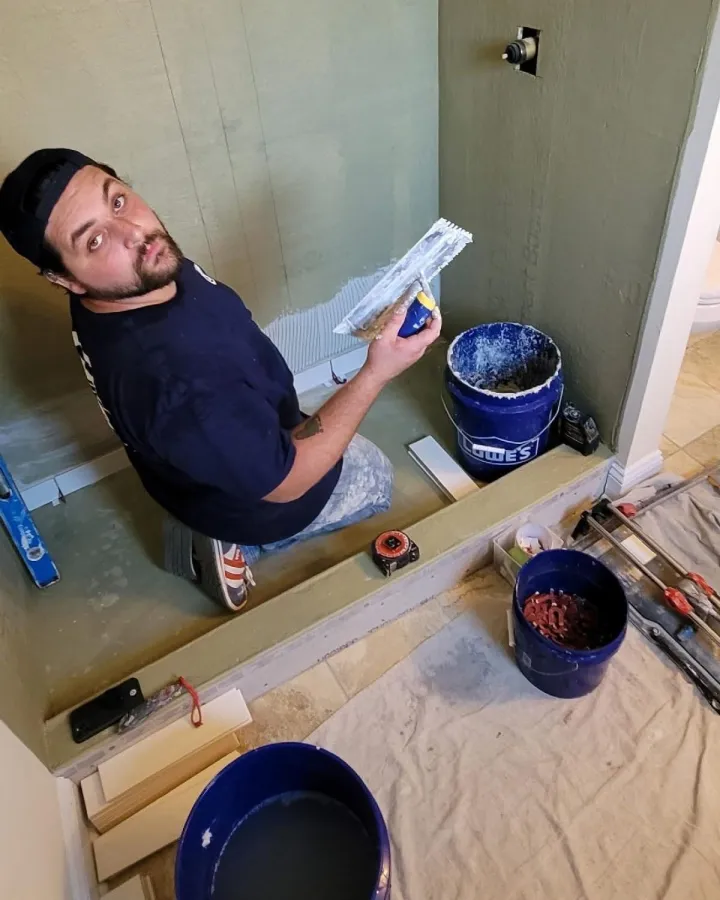 Brandon kneels in a shower and holds a notched trowel while preparing a wall for tile installation.