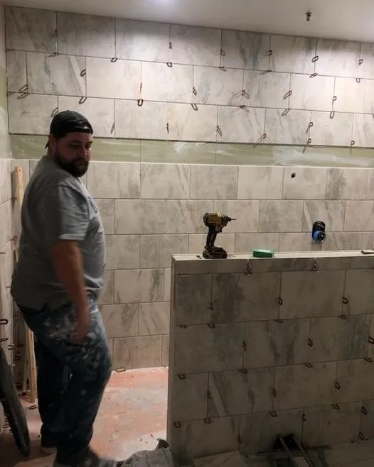 Brandon stands inside a partially tiled shower surrounded by large gray wall tiles and leveling clips.
