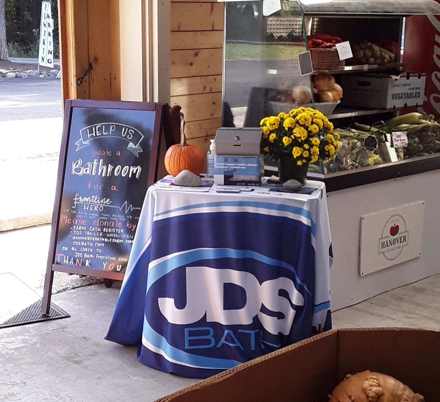 Placard sign for JDS fundraiser reads, “Help us update a bathroom for a frontline hero.” Next to it is a table with a JDS bath tablecloth, pumpkin, donation box, and flowers.