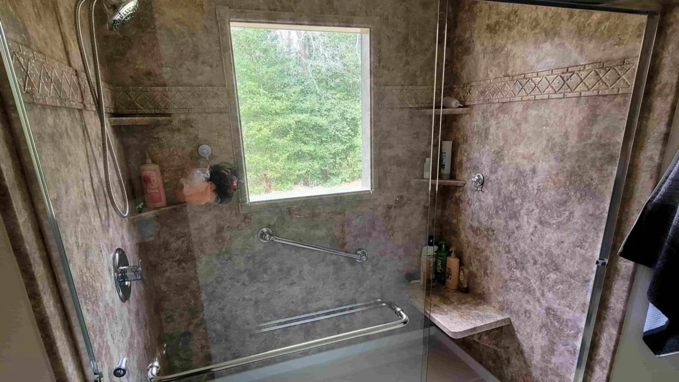 Beautiful accessible shower with sliding glass door and brown surround with window outlooking a green nature view