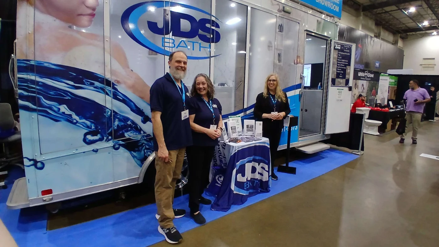 Paul and Vickie stand beside a blue and white JDS Bath mobile showroom at an indoor event.